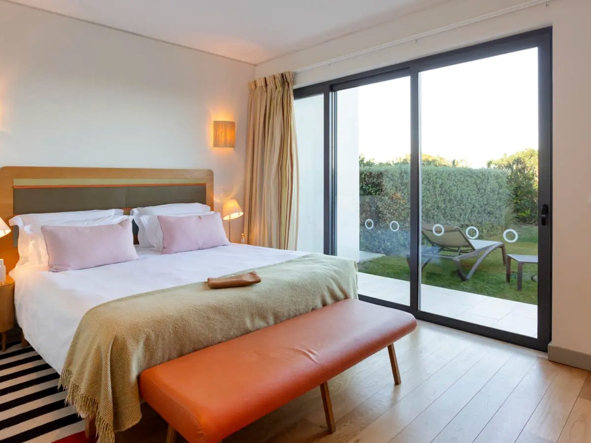 A bedroom with a double bed, an orange leather bench at its foot and sliding glass doors onto a hedged lawn with two sun loungers.