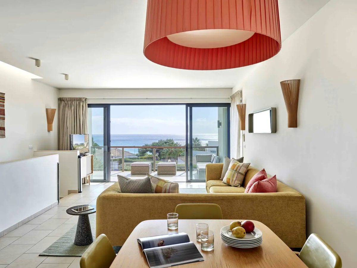 An open-plan living and dining room with a mustard sofa, a red drum pendant light, a wooden dining table and sliding doors onto a balcony with a sea view.