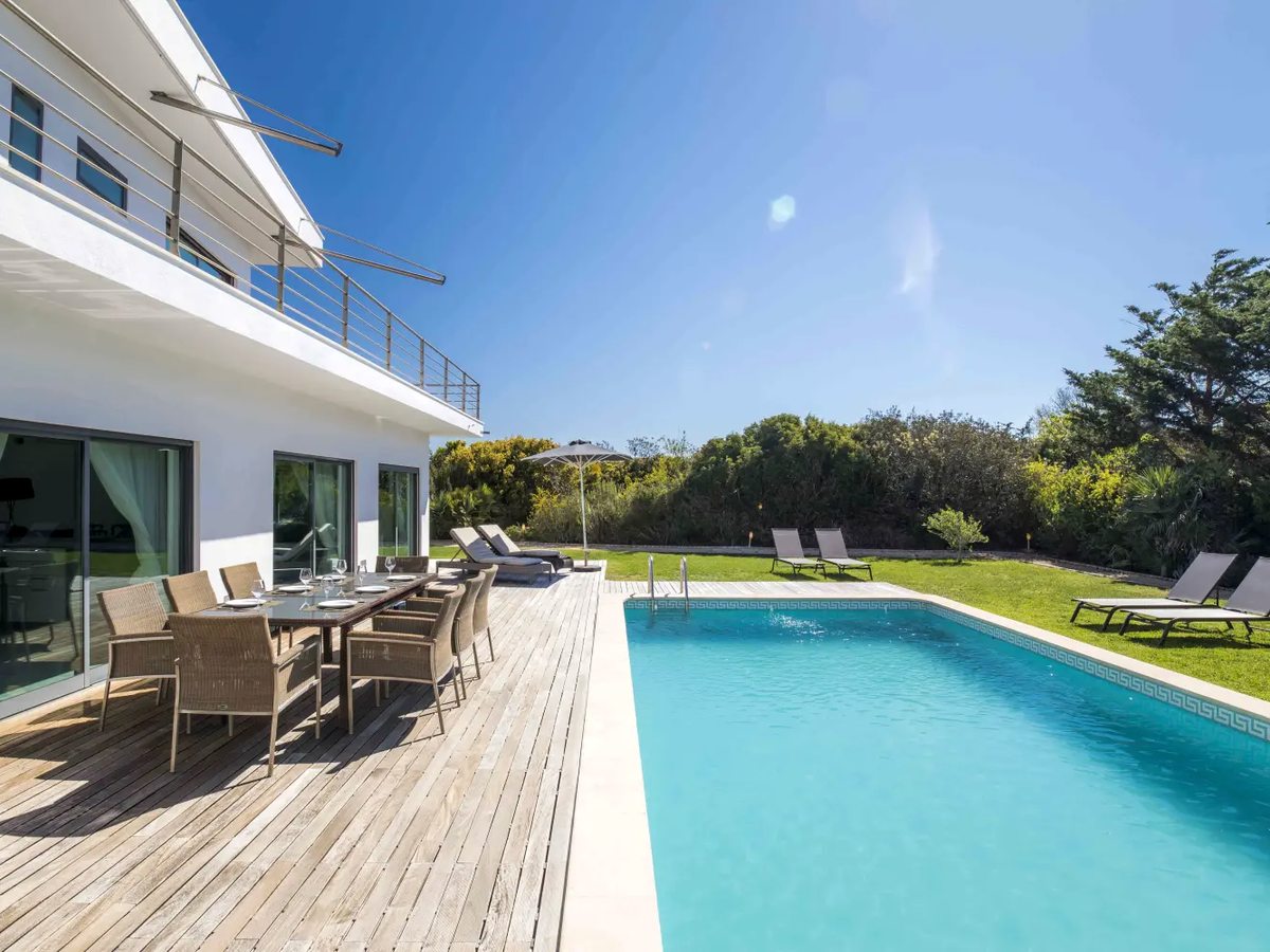 A rectangular pool beside a timber deck and lawn at a white flat-roofed villa, with an outdoor dining table, sun loungers and coastal scrub behind.