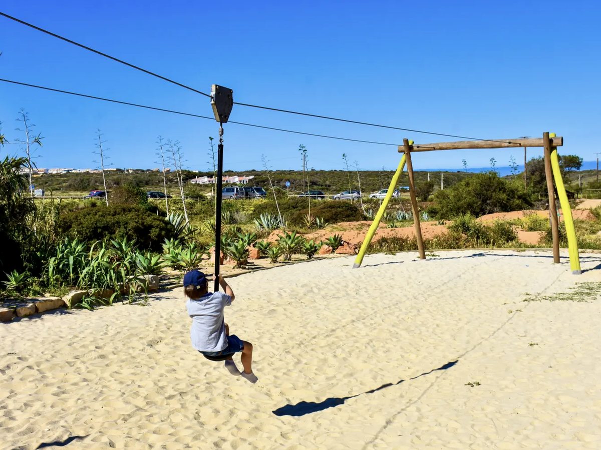 A small child in a cap riding a zip line seat across a sandy play area, with agaves, scrub and a timber swing frame behind.
