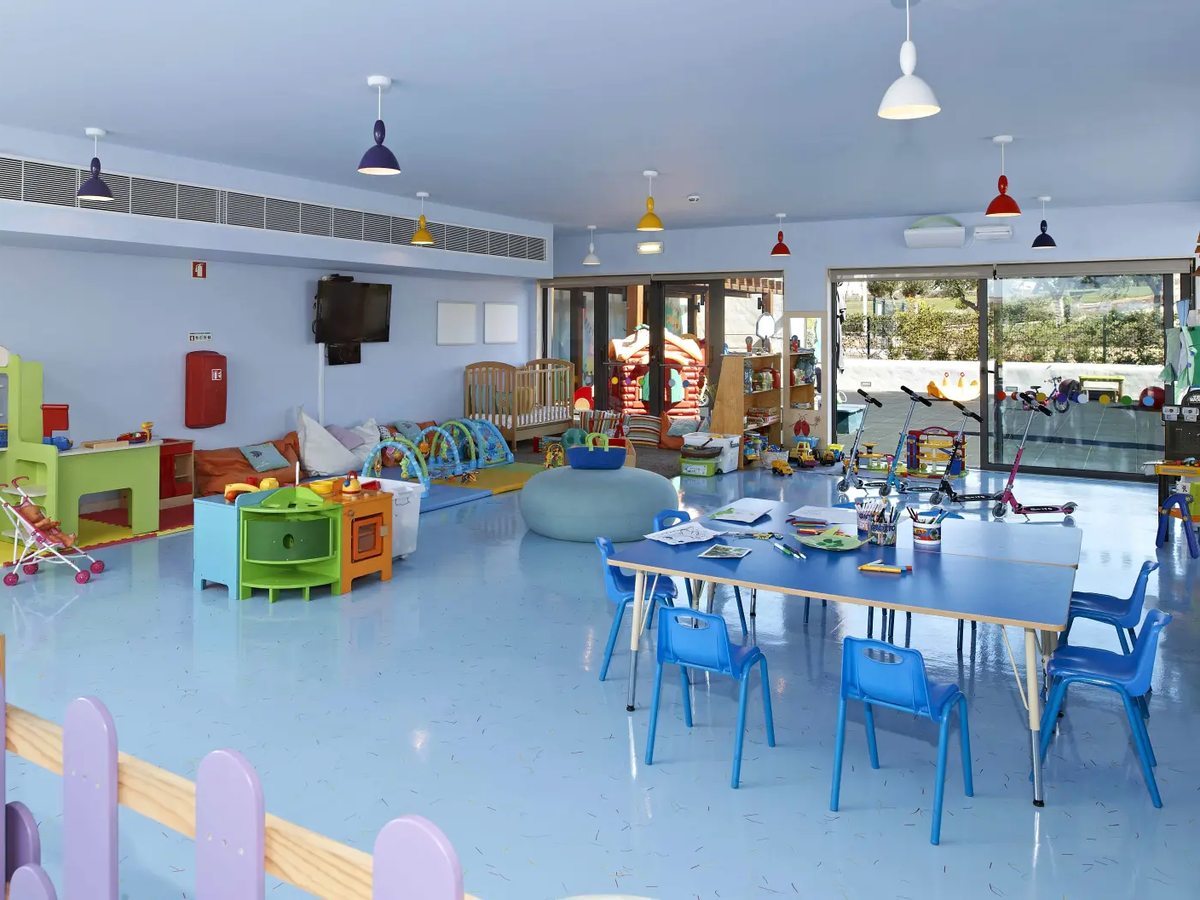 A pale blue playroom with a low blue table and child-sized chairs set out with drawing things, a wooden cot, a toy kitchen, soft play mats and scooters lined up by open glass doors.