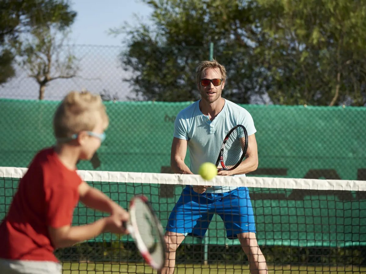 A coach at the net feeding a ball to a boy holding a racket on a tennis court backed by green windbreak netting and trees.
