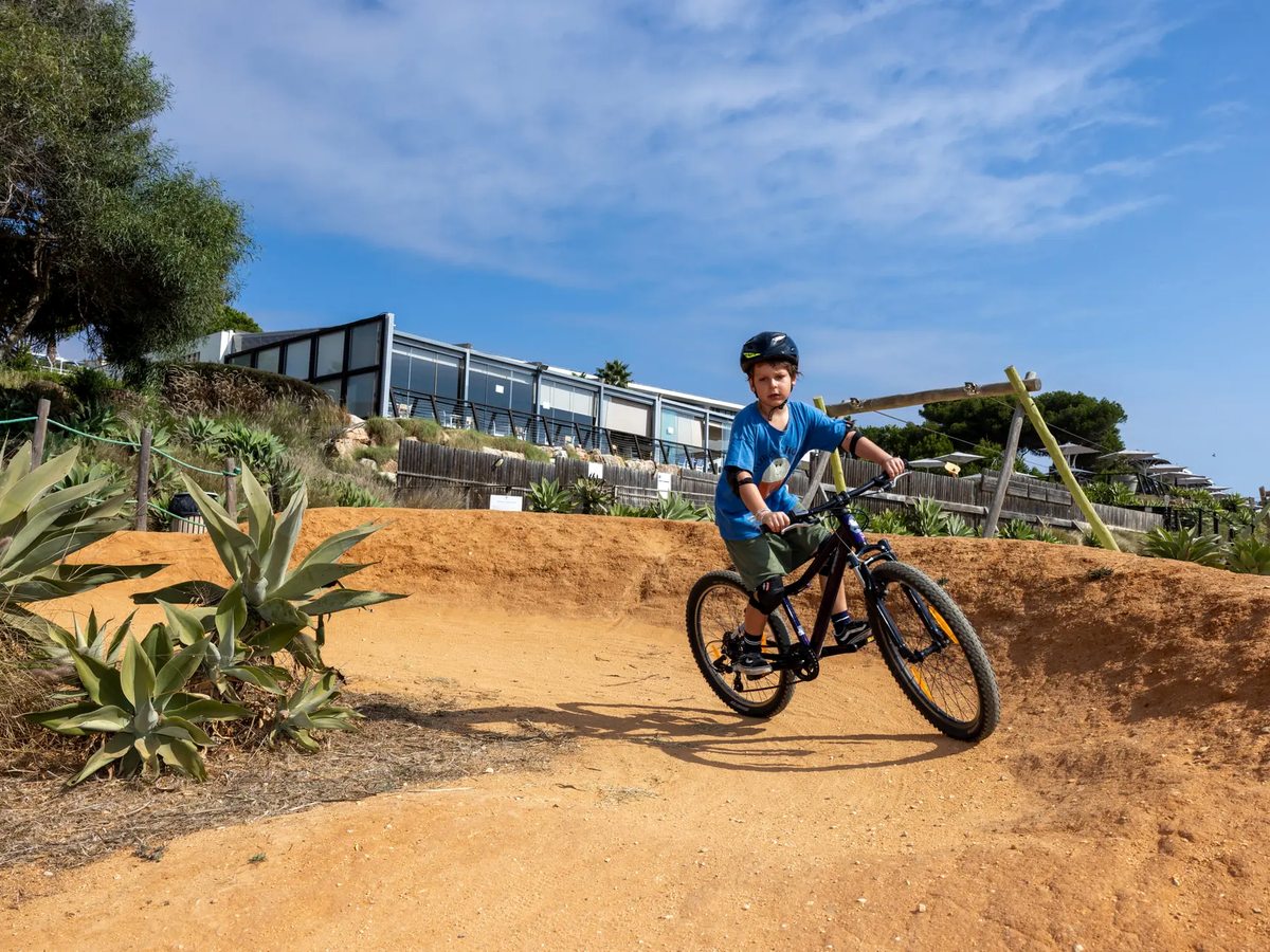 A boy in a helmet riding a mountain bike over the banked earth curves of a pump track, with a glazed resort building on the slope behind.