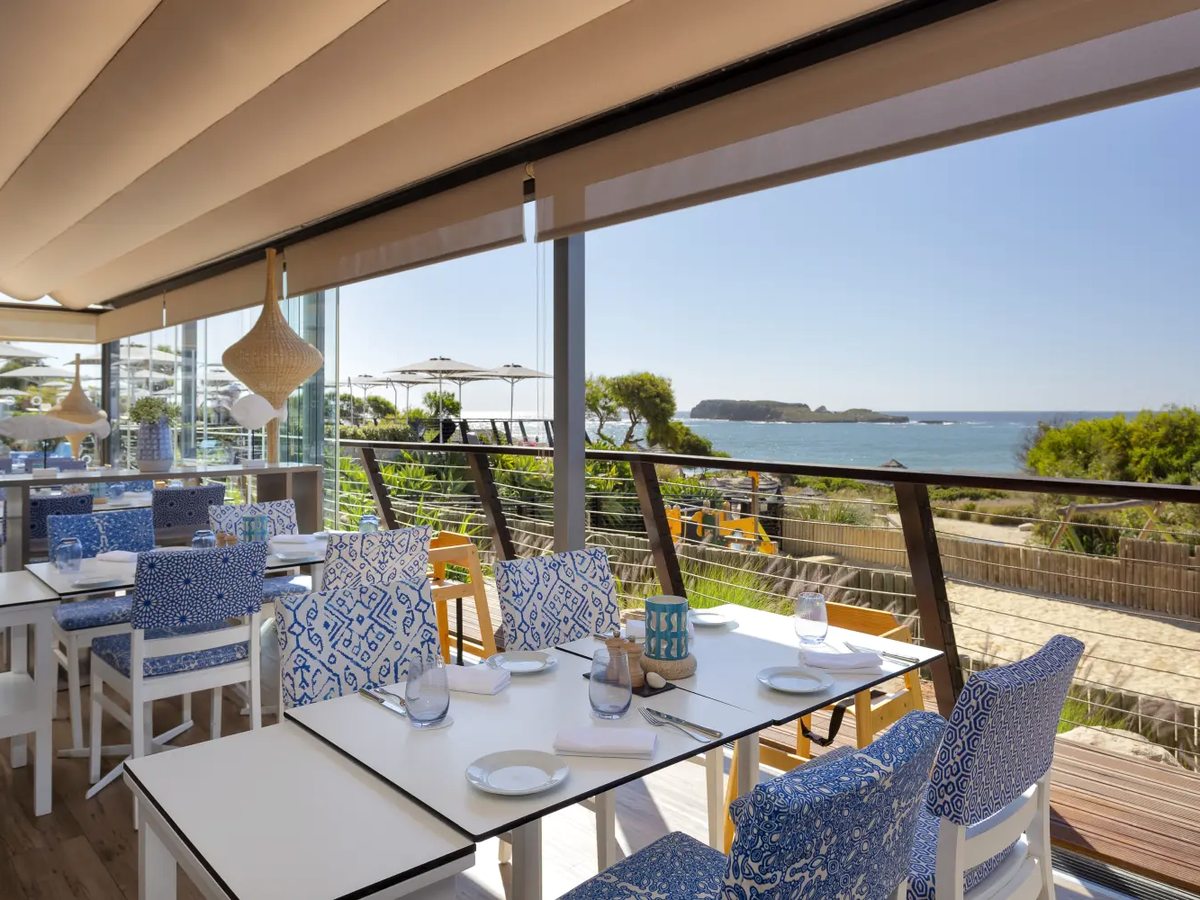 A covered restaurant terrace with white tables, blue patterned chairs and wooden high chairs, open to the dunes, the beach and an offshore island.