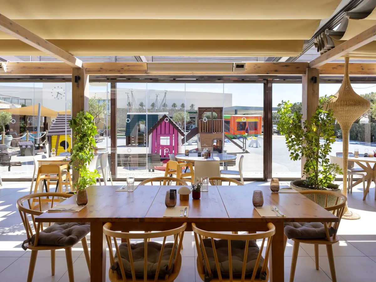 A restaurant with long wooden tables under a cream canopy, its glass wall open to a playground of pink and blue wooden play houses and a climbing frame.
