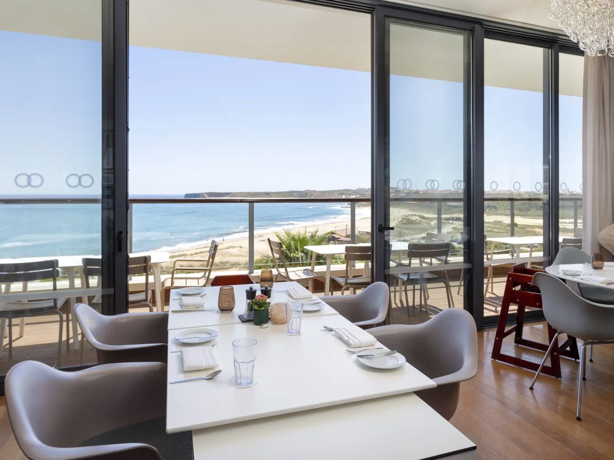 A dining room with white tables and grey chairs behind full-height glass doors, a wooden high chair at one table and the sandy bay and headland beyond.