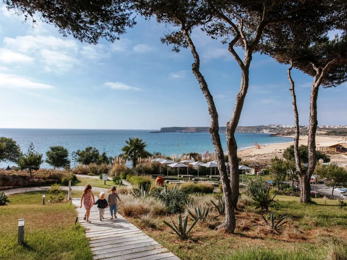 A timber boardwalk running down through grass, agaves and umbrella pines to a sandy beach and blue bay, with a woman and two small children walking up it.