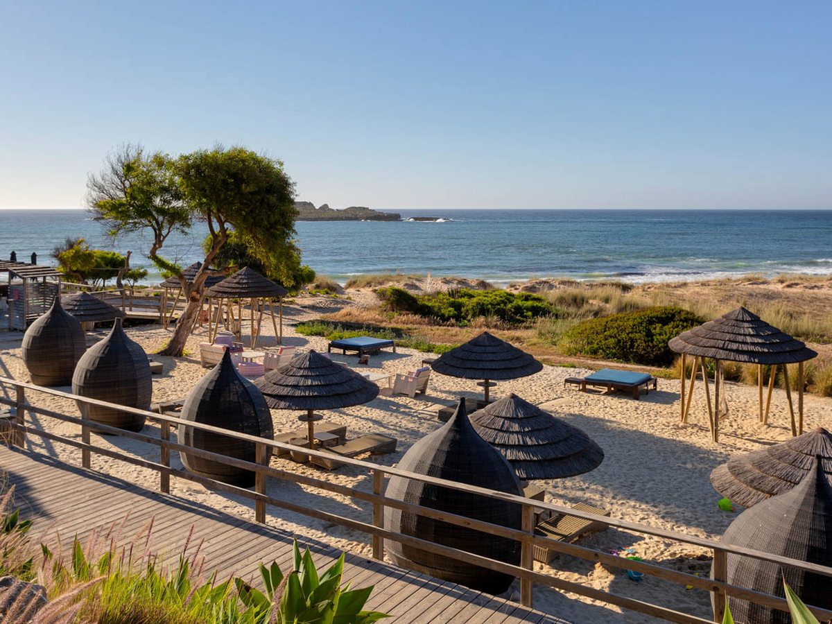 Woven pod chairs and thatched parasols on the sand beside a timber walkway, with dunes, the bay and an offshore rock beyond.