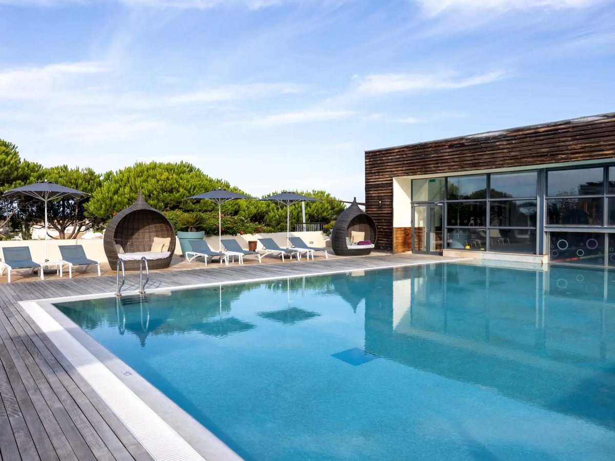 A rectangular outdoor pool edged with timber decking, sun loungers and two round wicker shade pods, running up to a glazed timber-clad building.