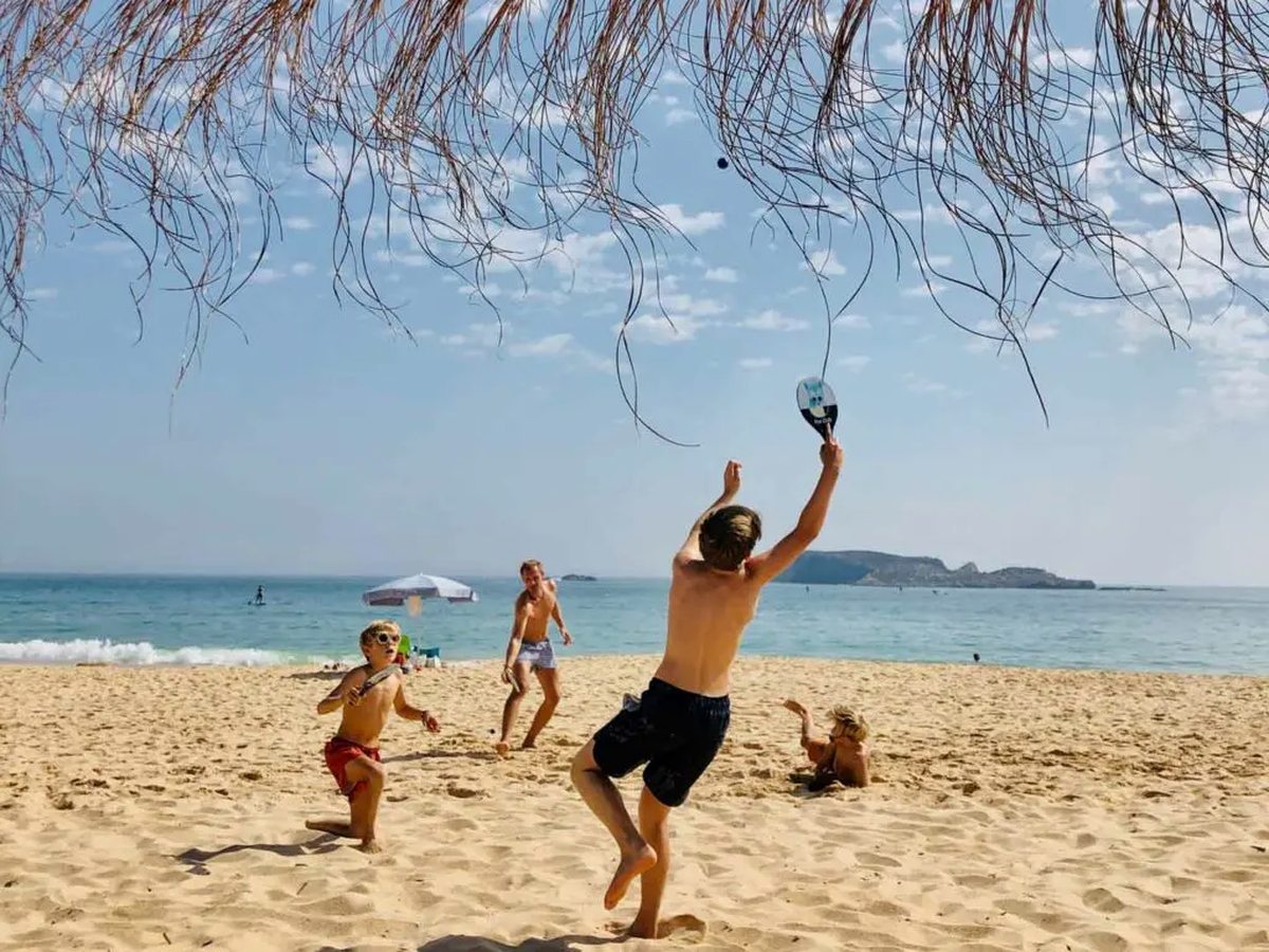 Children and adults playing bat and ball on the sand below a thatched shade, with the sea and an offshore island behind.