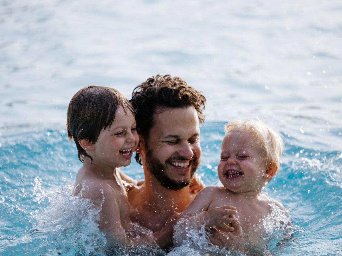 A man in a sunlit pool holding two laughing children as they splash in the water.