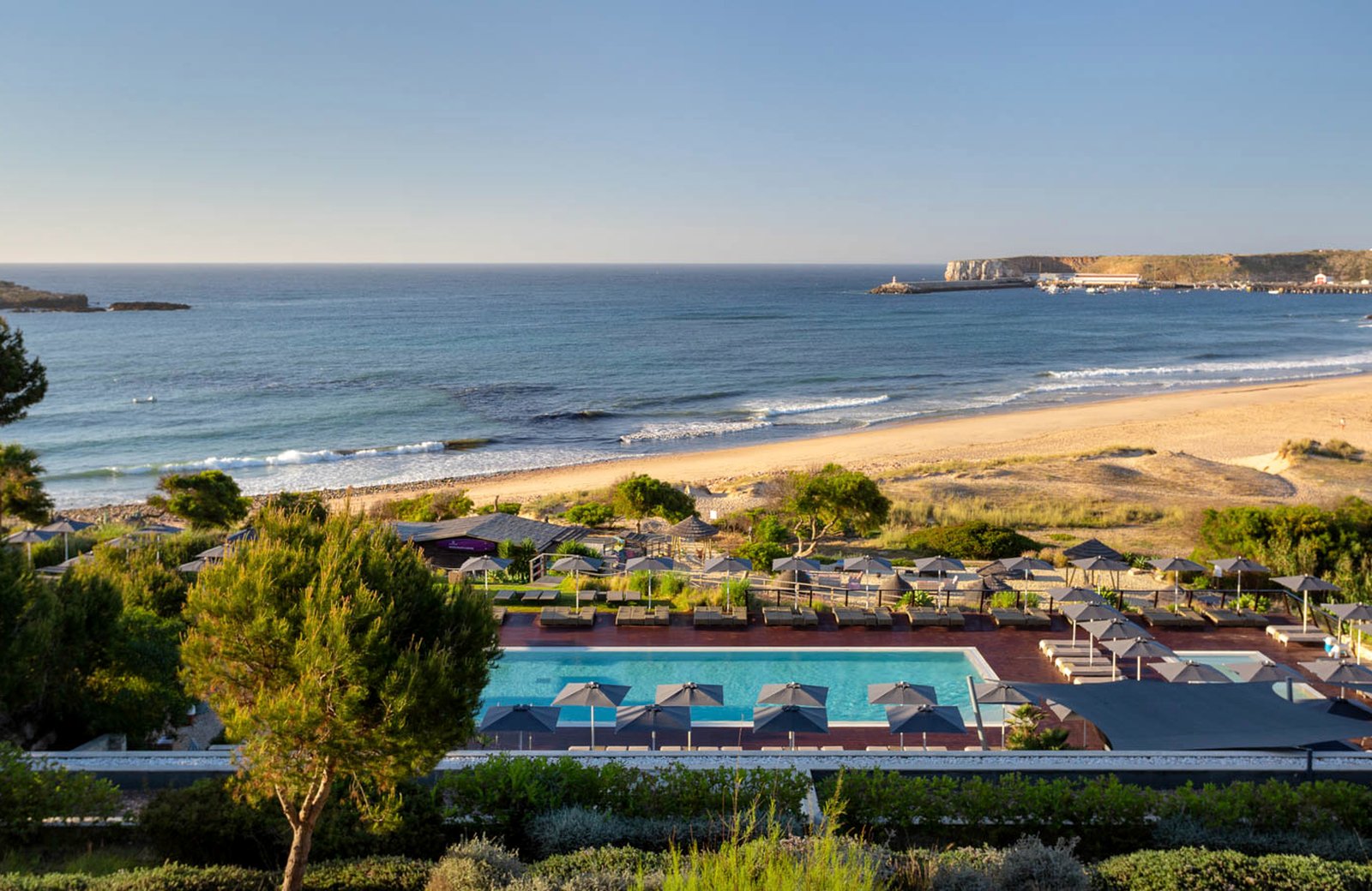 The resort's beachside pool and rows of sun loungers under grey parasols on a terrace above the wide sandy sweep of Martinhal beach, with the Sagres headland and harbour across the bay.
