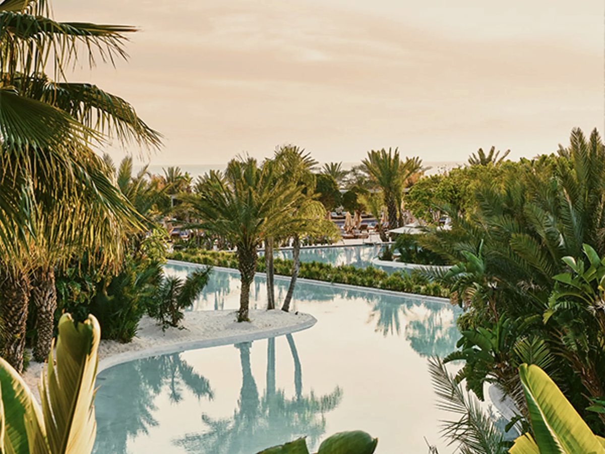 A lagoon-shaped pool curving between planted beds and palms on white sand, with parasols and the sea in the distance.