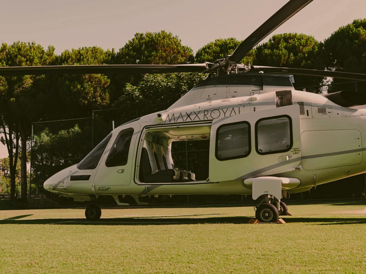 A white Maxx Royal helicopter standing on a grass strip with a line of pine trees behind it.