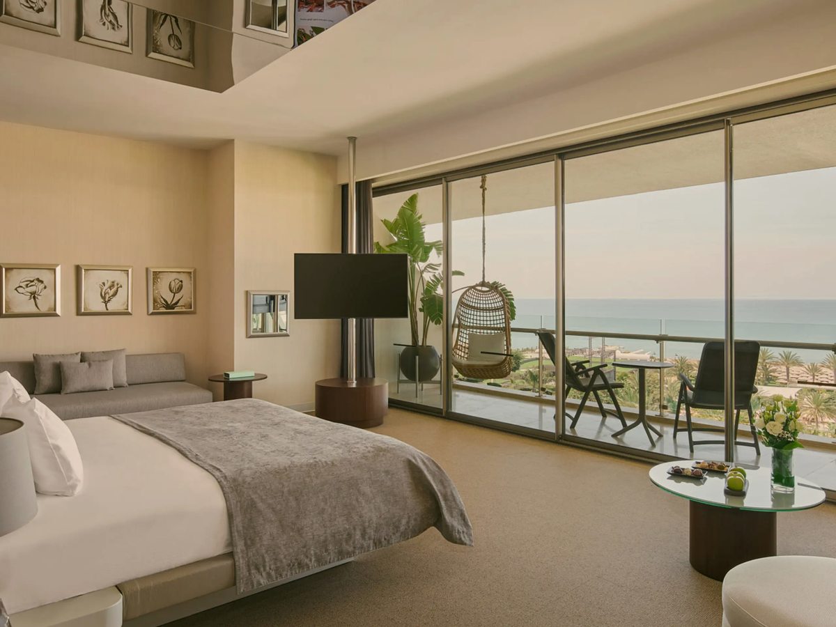 A suite bedroom with a large bed, framed prints, a hanging chair by the window and a full-height glass wall onto a balcony with the sea beyond.