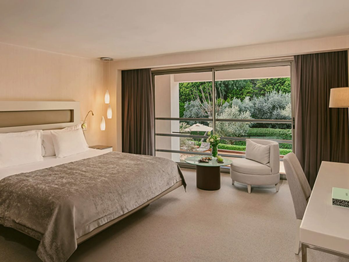 A suite bedroom with a large bed, a desk and an armchair, and a full-height window onto a planted garden.