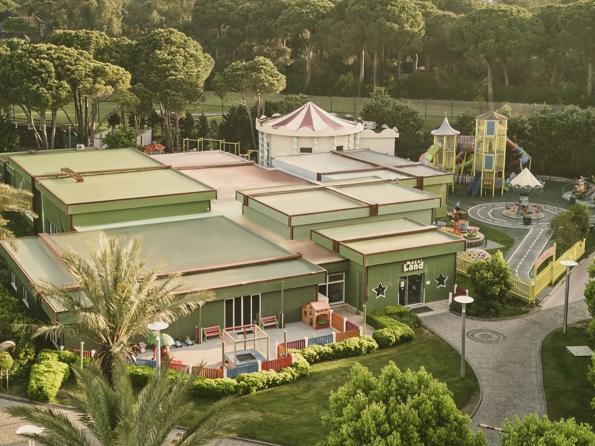 An aerial view of low green-roofed buildings with a striped carousel canopy, climbing frames and slides and a small road circuit, ringed by palms and pines.
