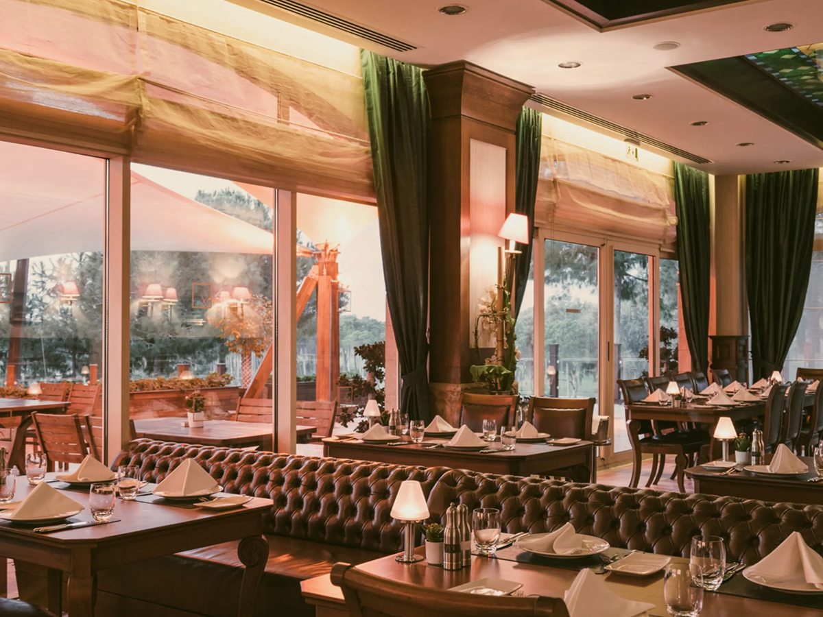 A formal restaurant with dark tufted leather banquettes, laid tables with white napkins and small lamps, tall windows and green drapes.