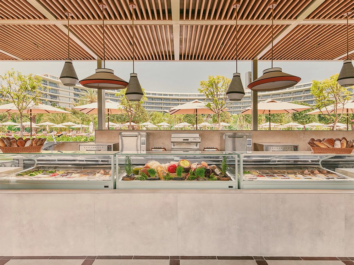 An open counter under a slatted timber roof with chilled displays of fish, vegetables and bread, the curved resort building and rows of parasols behind.