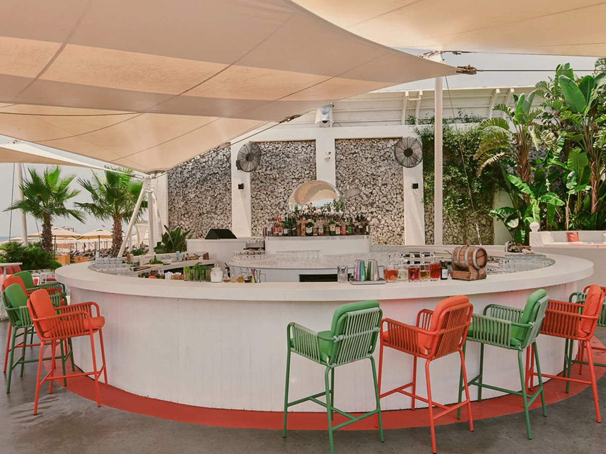 A circular white bar with coral and green woven stools under a white shade sail, palms and greenery around it.