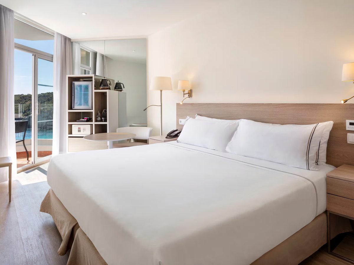 Hotel bedroom with a king bed, a pale wood headboard and bedside lamps, beside a shelving unit with a television, kettle and safe, and balcony doors.
