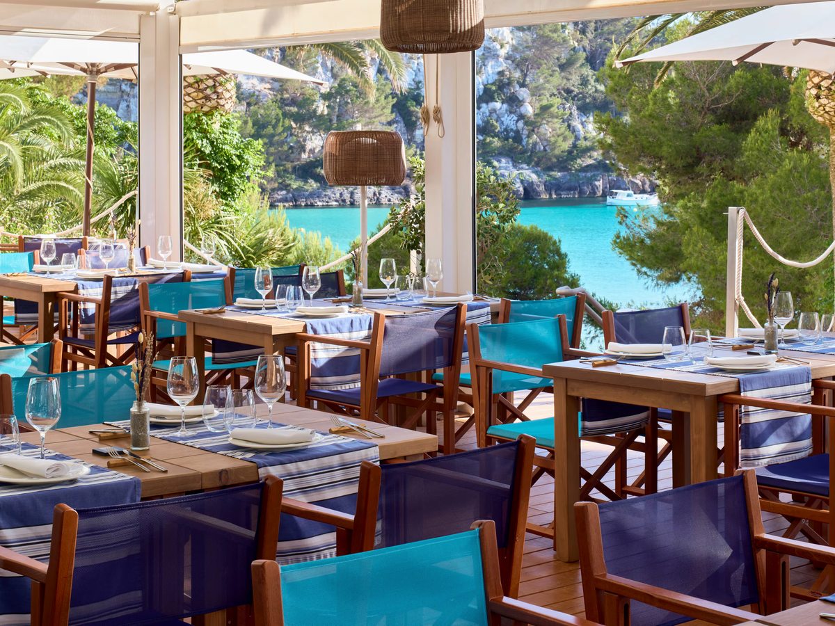 Outdoor restaurant terrace with wooden tables, blue and turquoise director's chairs and laid place settings, with pines and turquoise water beyond.