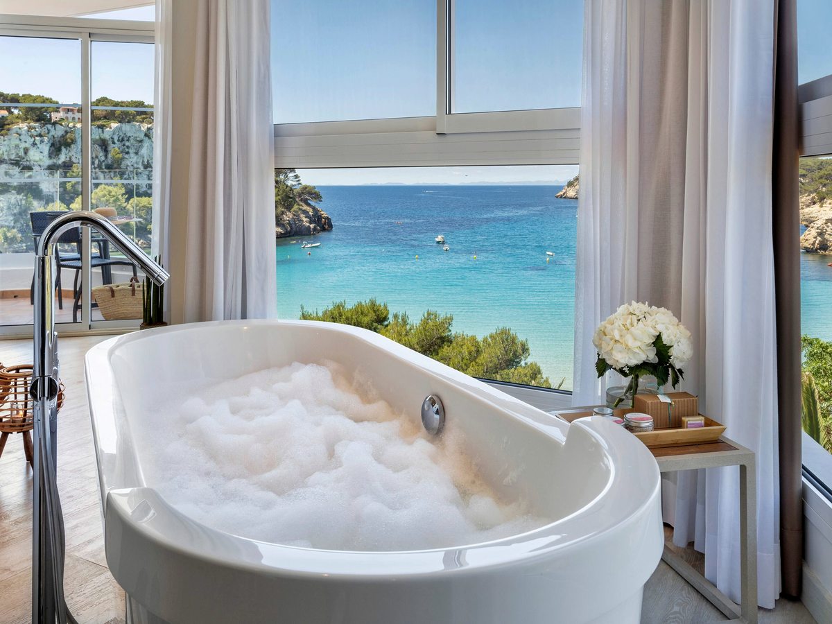 A freestanding white bath filled with bubbles beside full-height windows, with a wooden bath tray, toiletries and white flowers alongside.