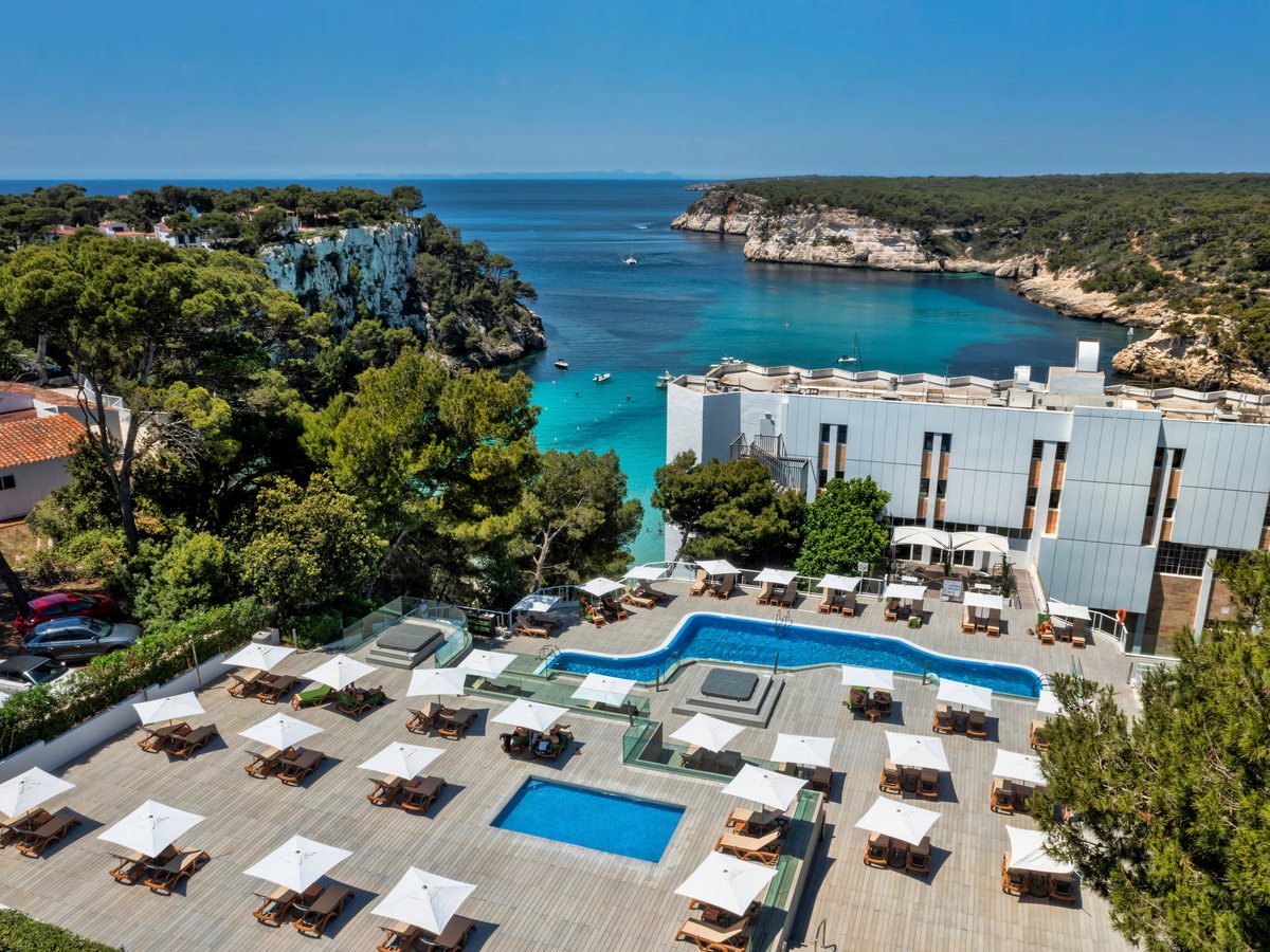 Curved outdoor pool and a smaller square pool on a wide timber deck of sun loungers and white parasols, with the hotel behind and the turquoise cove beyond.