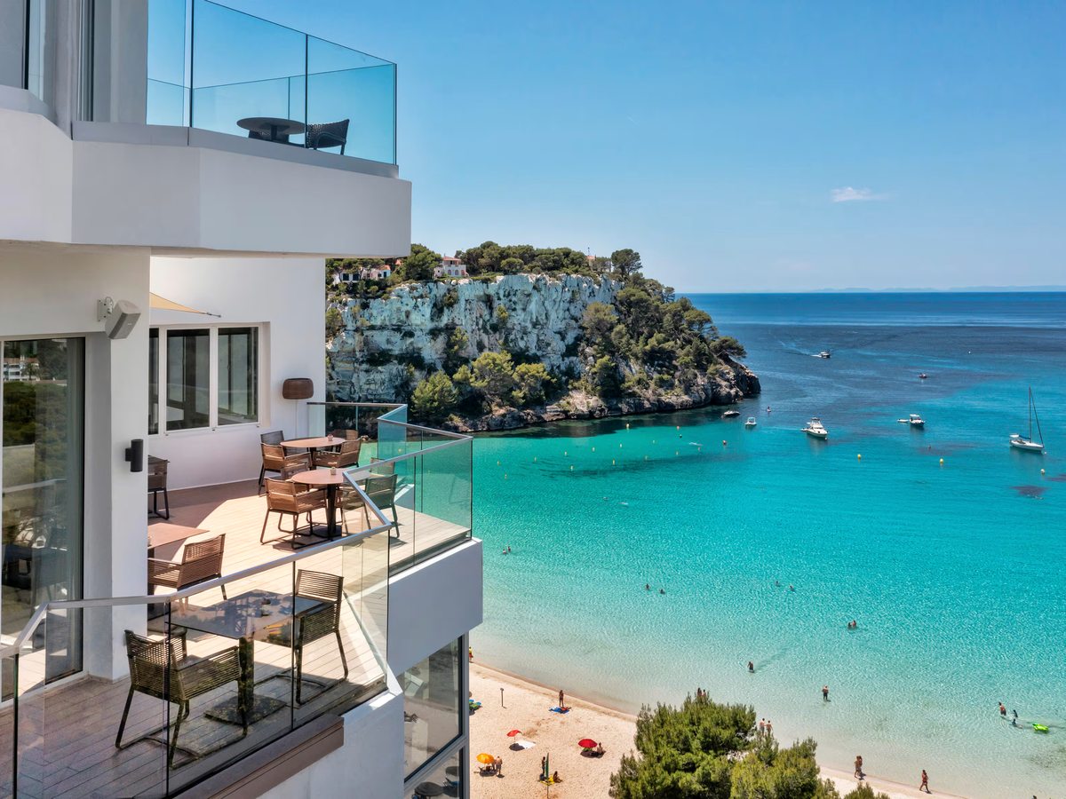 White hotel balconies with glass balustrades and terrace tables, above the sandy beach and turquoise water of a cove ringed by pine-topped cliffs.
