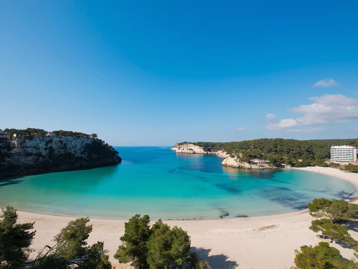 Horseshoe cove of pale sand and clear turquoise water, enclosed by pine-topped limestone cliffs under a blue sky.