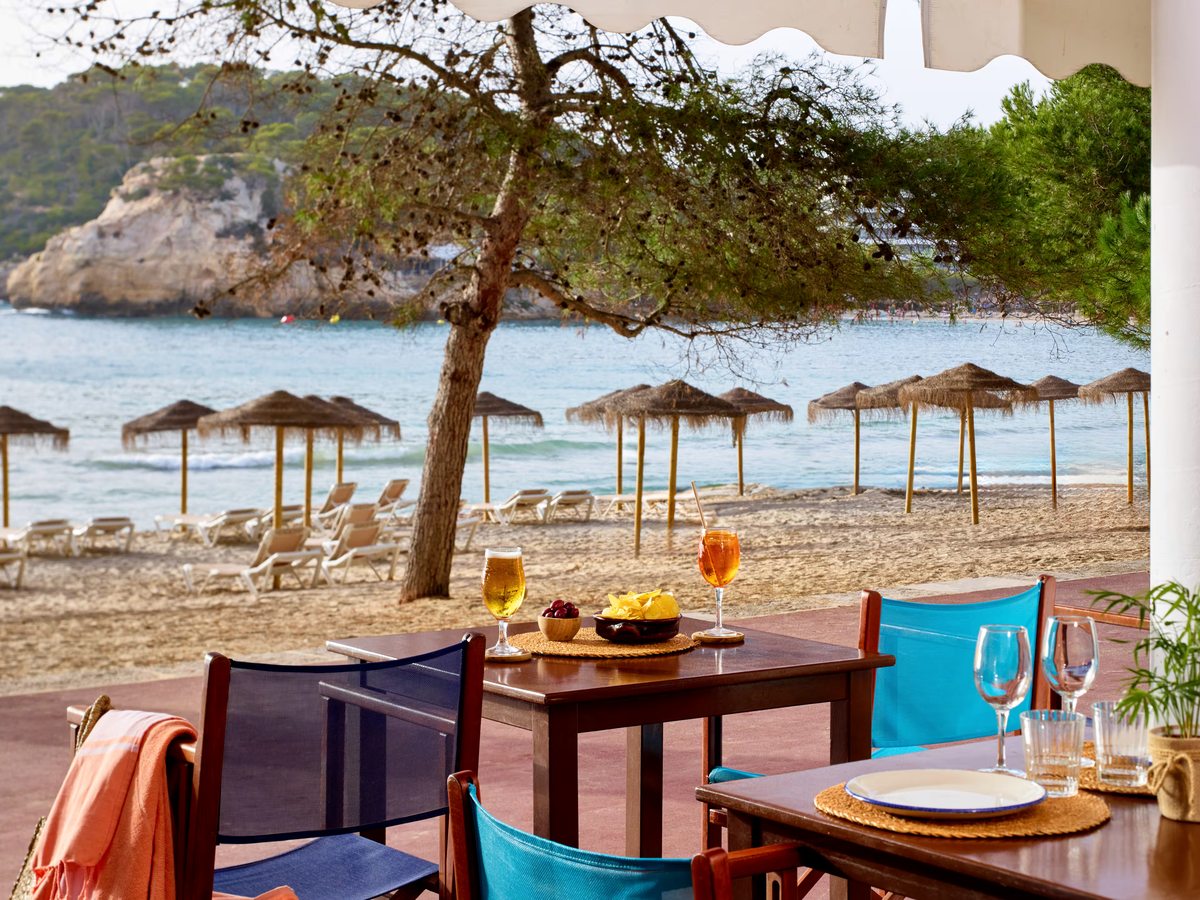 Wooden tables and blue canvas chairs under a shaded terrace at the edge of the sand, with straw parasols, sun loungers and the cove behind.