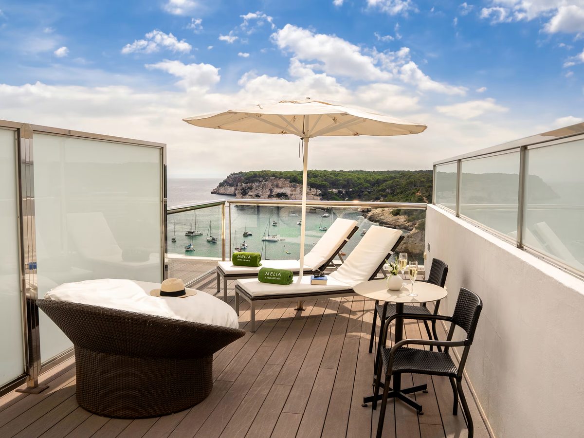 Decked private terrace with two sun loungers, a parasol, a round table and a large woven daybed, above a cove of moored yachts and pine-topped cliffs.