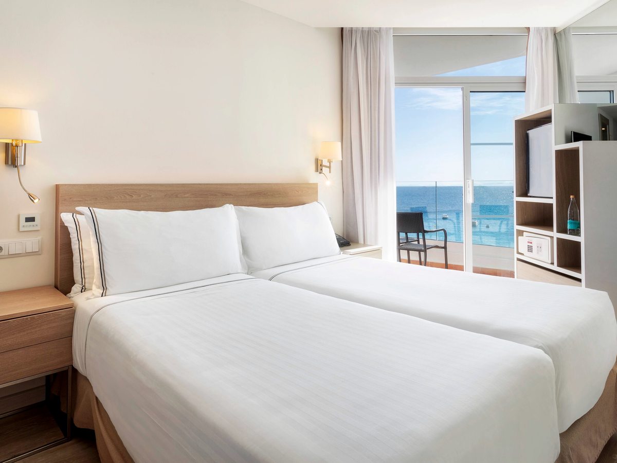 Bedroom with two single beds pushed together under a wooden headboard, wall lamps either side and a furnished balcony through glass doors.
