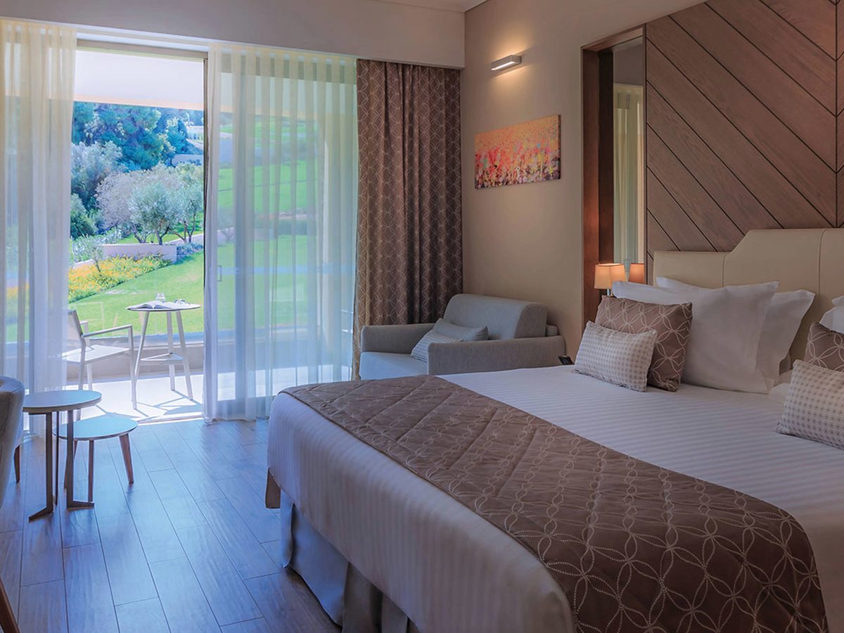 A double bed with a patterned runner, a small sofa and a wall mirror, with glass doors open to a balcony table and a lawn beyond.