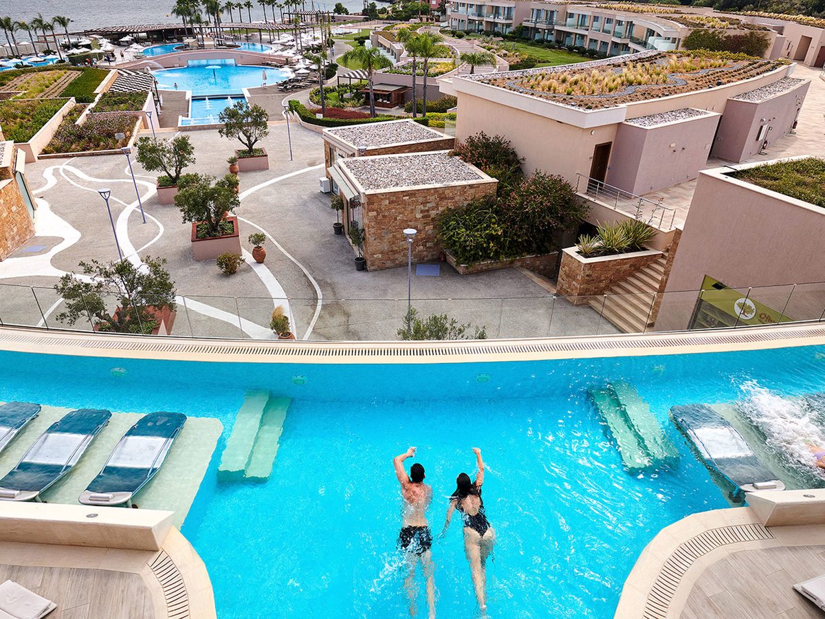 A curved pool with padded loungers set in the shallow water and two swimmers, with terraced guest buildings and planting behind.