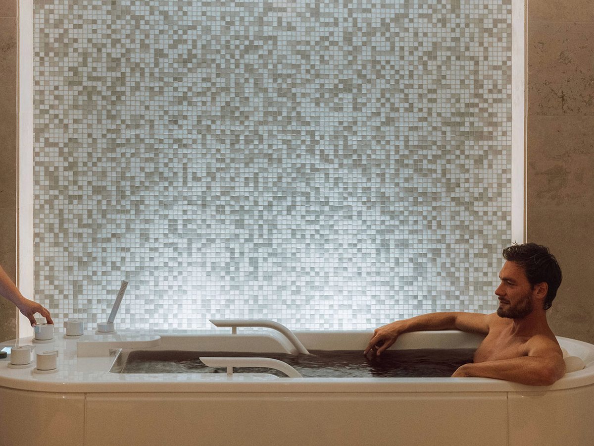 A man lying back in a white hydro bath in front of a lit mosaic wall, with a therapist reaching in at the side.