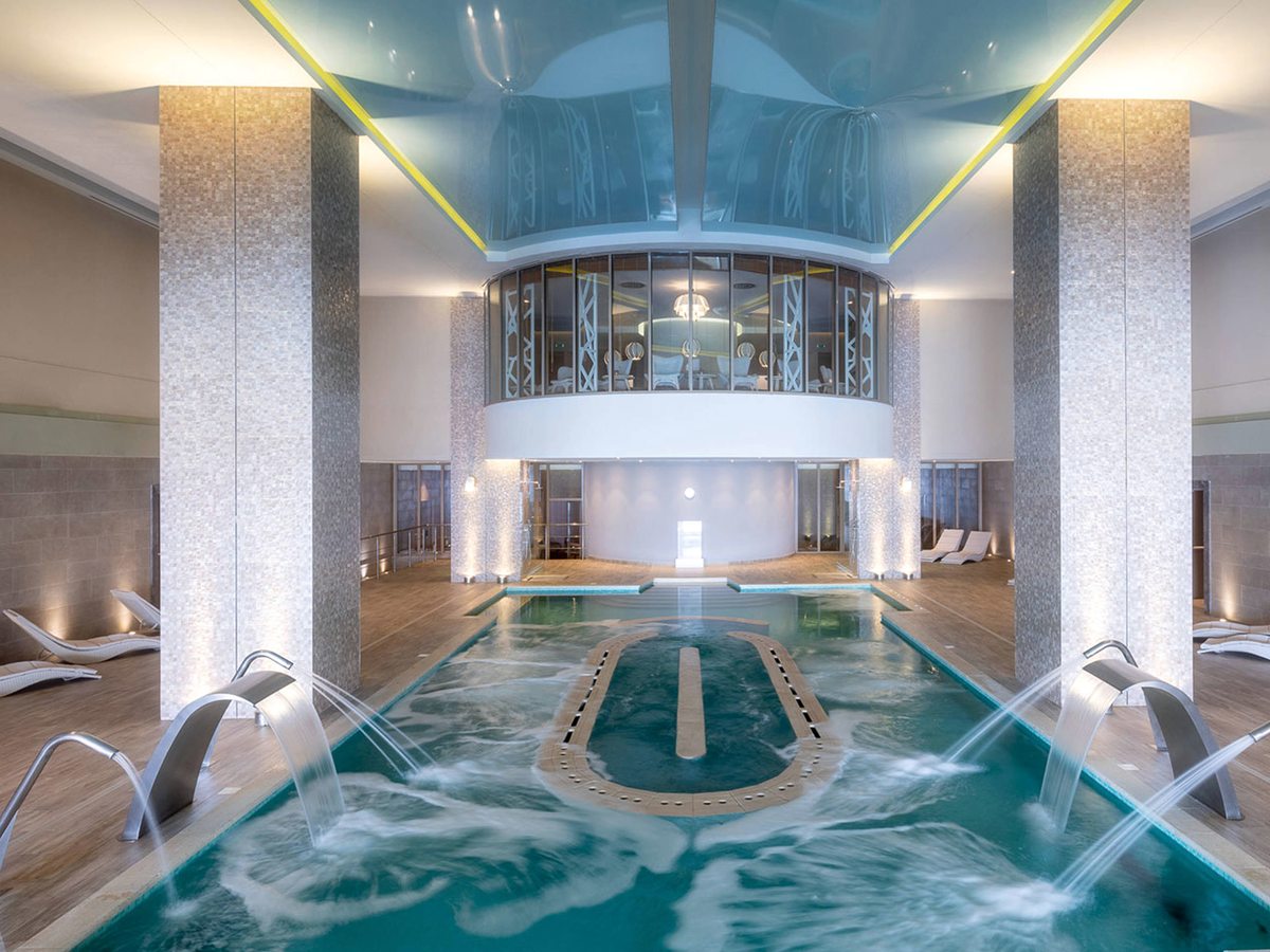 A long indoor pool with arcing water jets along both sides and a bubbling section in the middle, under a curved ceiling between tall mosaic columns.