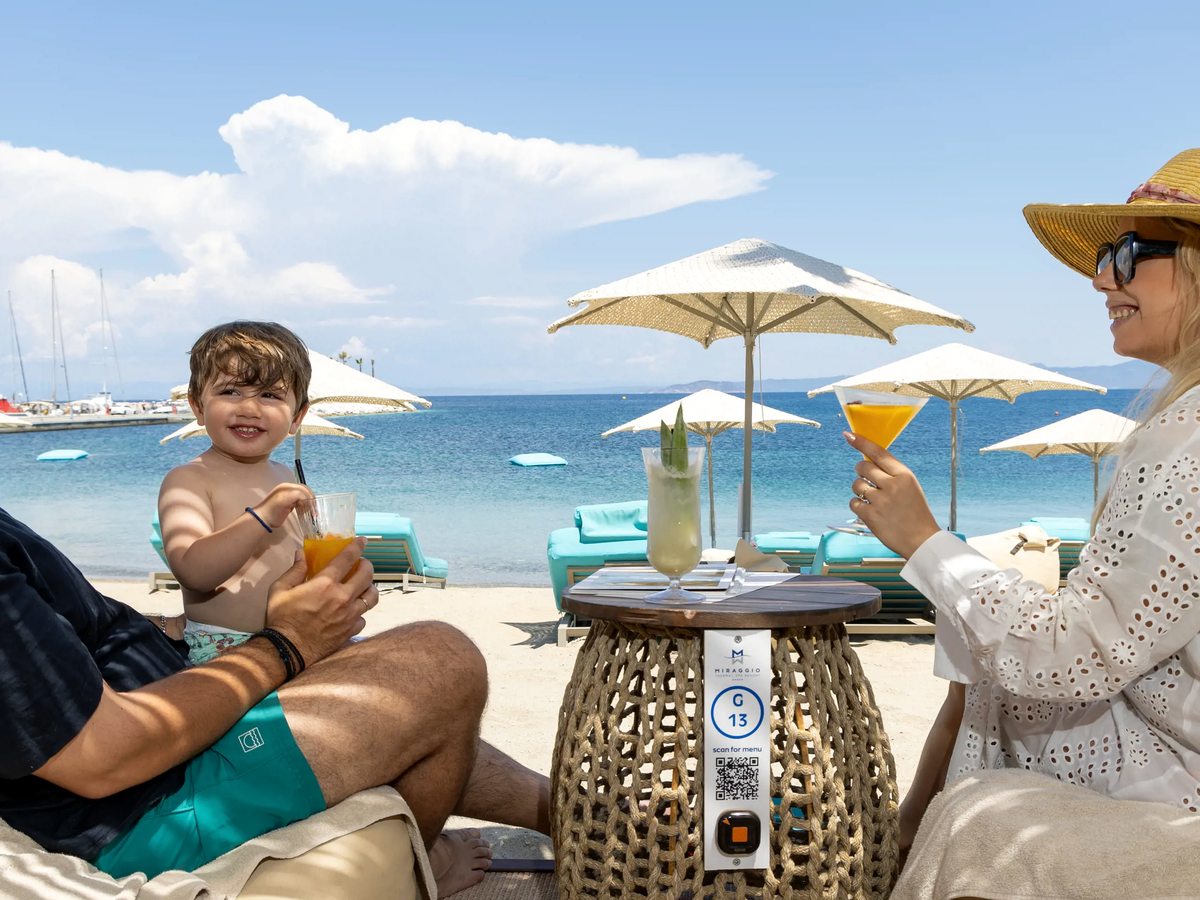 A man holding a toddler with a drink and a woman raising a glass at a small table on the beach, with woven straw parasols and the sea behind.
