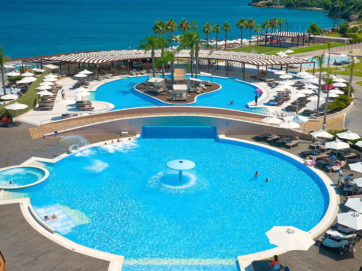 A large circular pool with a central fountain and a second curved pool alongside, ringed by sun loungers and white umbrellas, with palms and the sea behind.