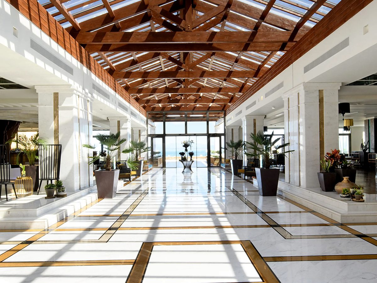 A long lobby with a pitched timber ceiling, white columns, a polished marble floor inlaid with dark bands, potted plants and glass doors at the far end.