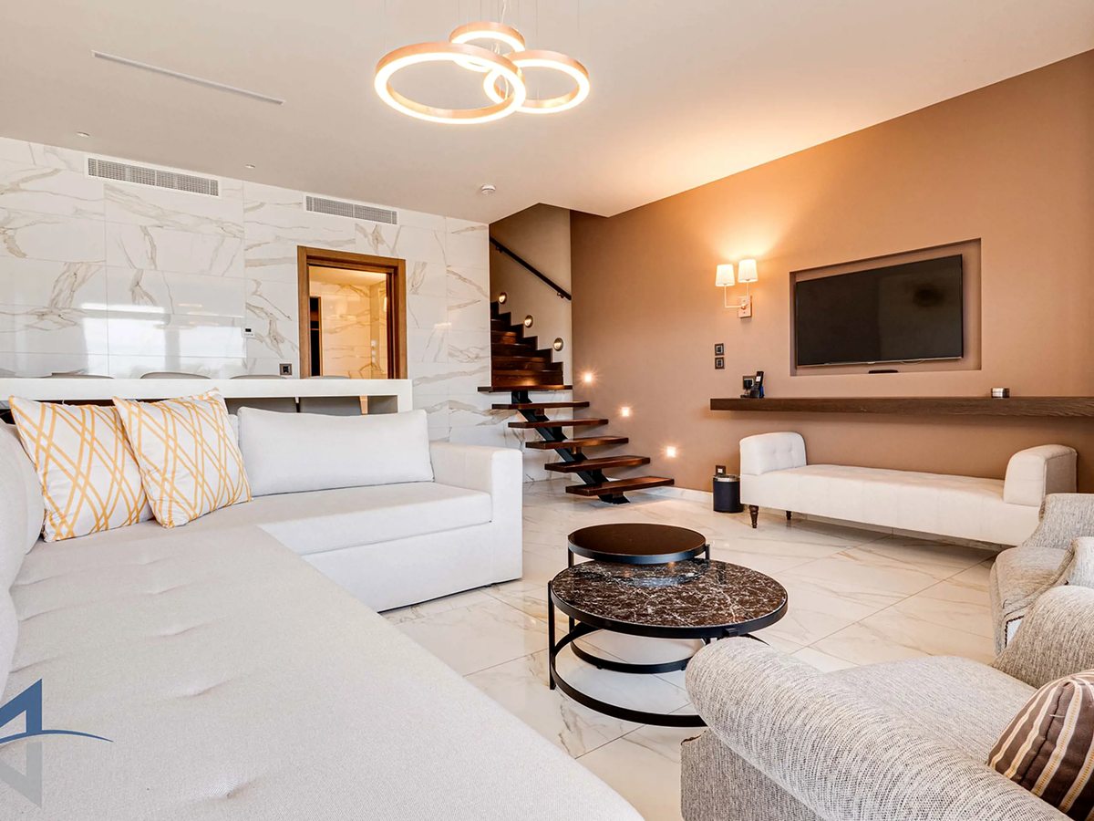 A living room with white sofas, a round coffee table, a wall-mounted television and an open dark-wood staircase rising to the upper floor.