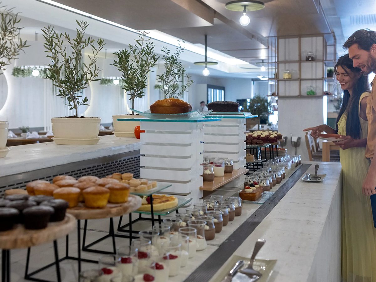 A tiered white counter of small cakes, tarts and pots of dessert, with olive trees in white planters behind and two guests choosing.