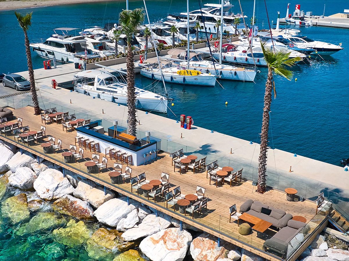 A timber quayside terrace lined with dining tables, chairs and a bar counter, with motor yachts and sailing boats moored along the marina beyond.