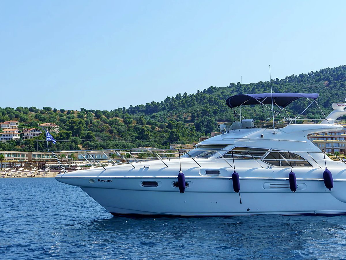 A white motor yacht on calm water, with a wooded headland, a village and resort buildings on the shore behind.