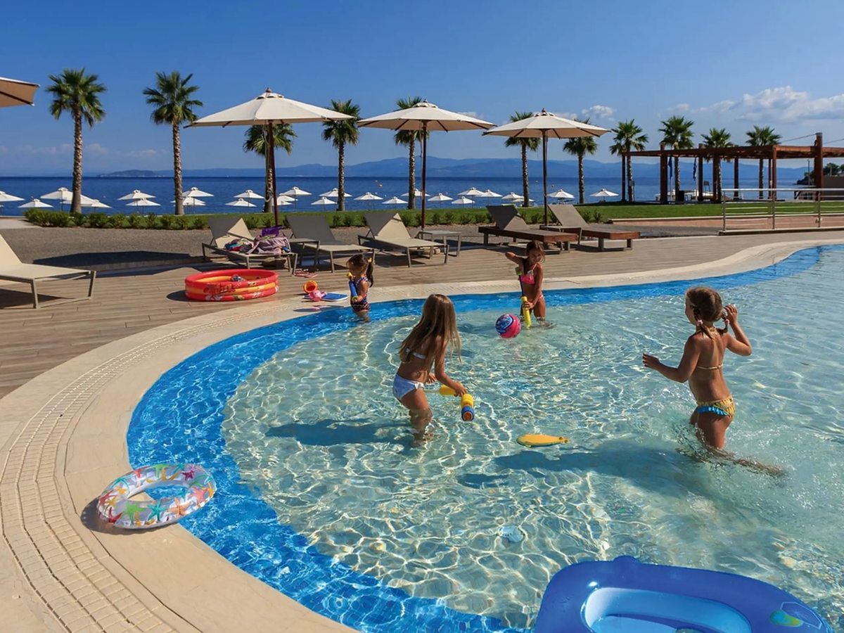 Children playing in a curved shallow pool with inflatable rings on the deck, sun loungers and umbrellas behind and the sea beyond.