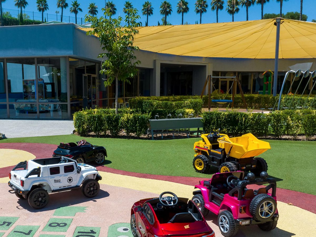 Child-sized electric cars parked on a coloured outdoor track beside a lawn, with hopscotch markings and the Kids Planet building under a yellow canopy behind.