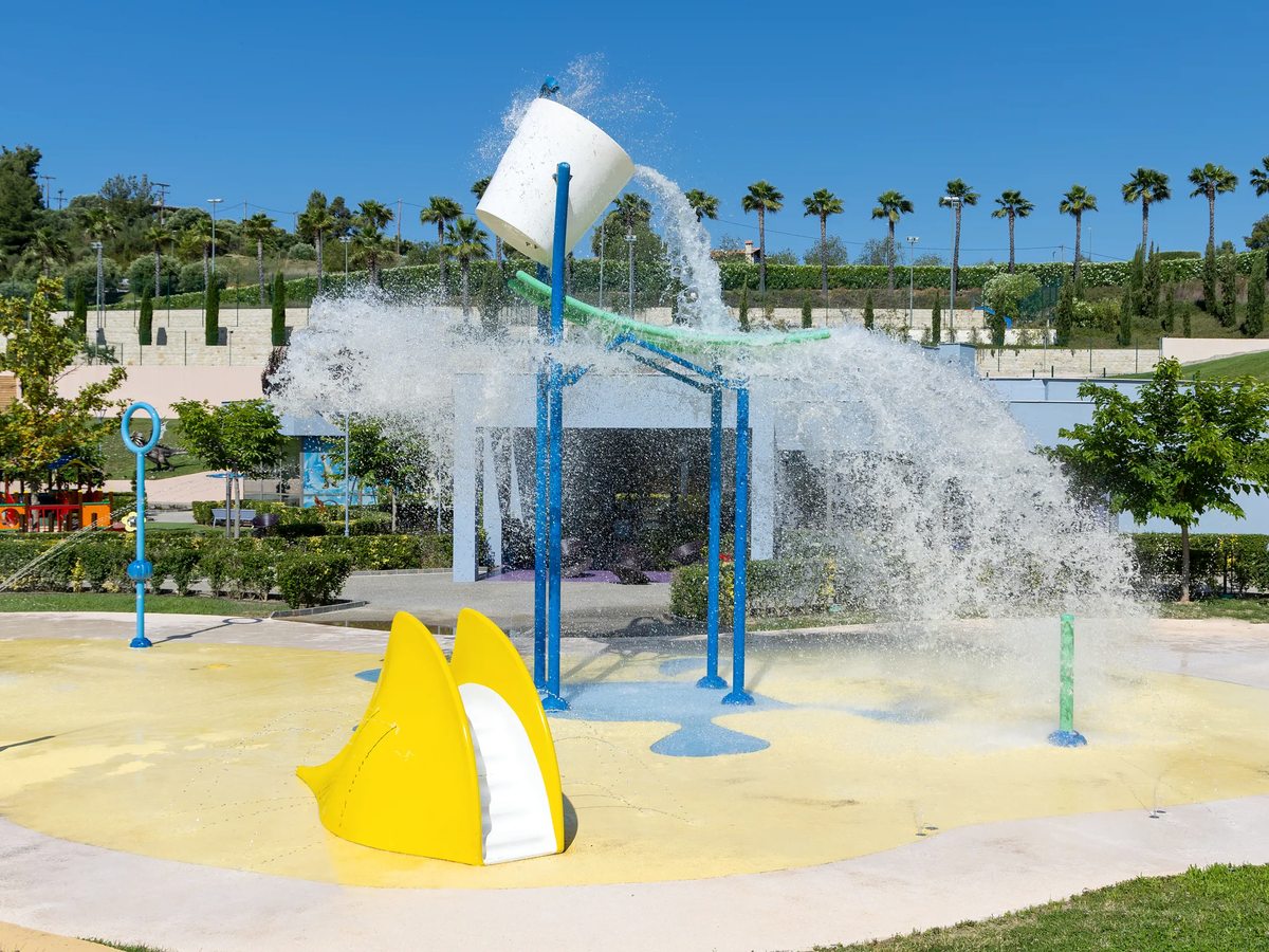 A splash pad with a tipping bucket pouring water over jets and a yellow slide, with palm trees and lawns behind.