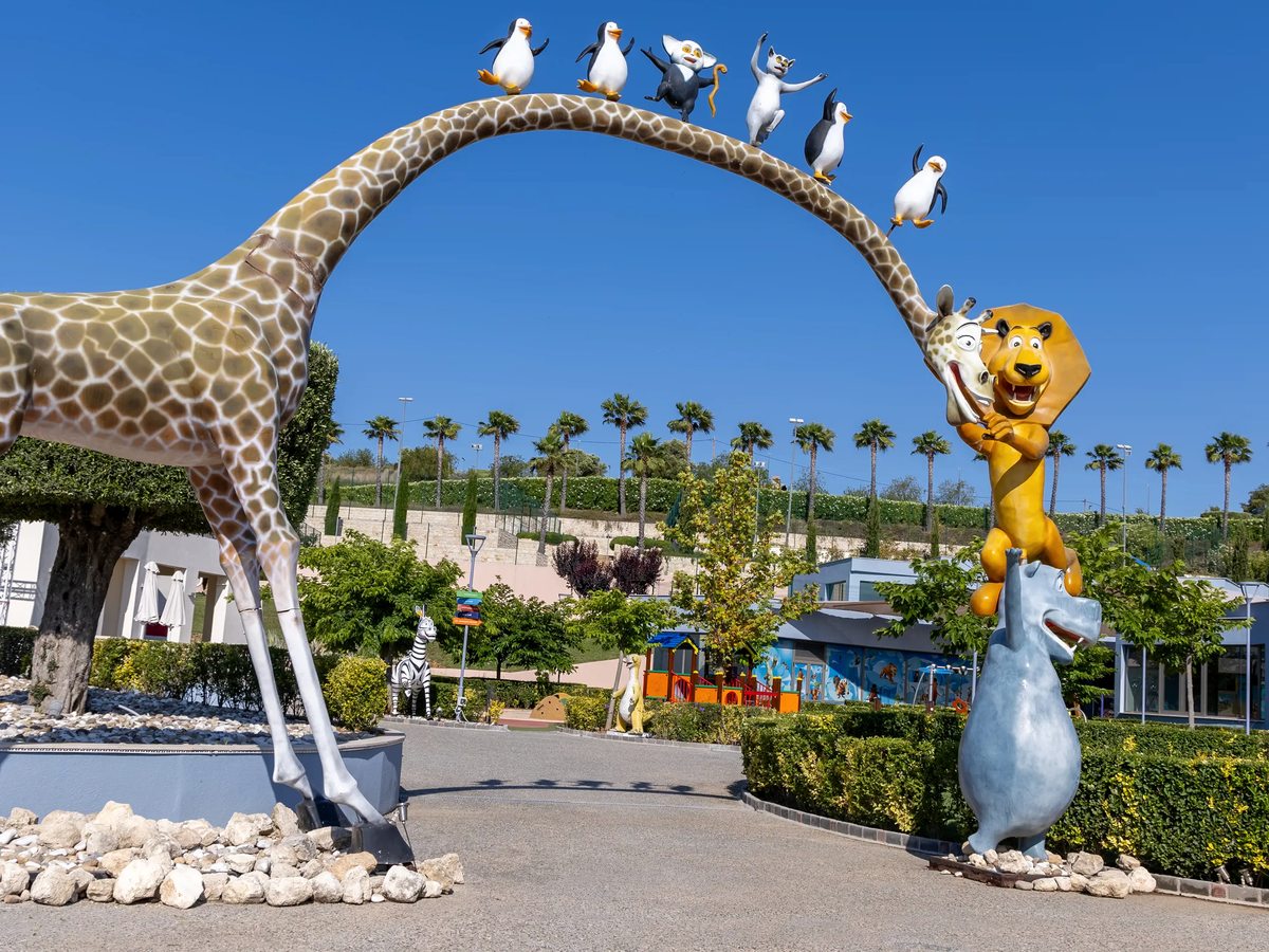 A giant giraffe figure arching over a path with penguin figures along its neck and a lion figure standing beside it, with palms and lawns behind.