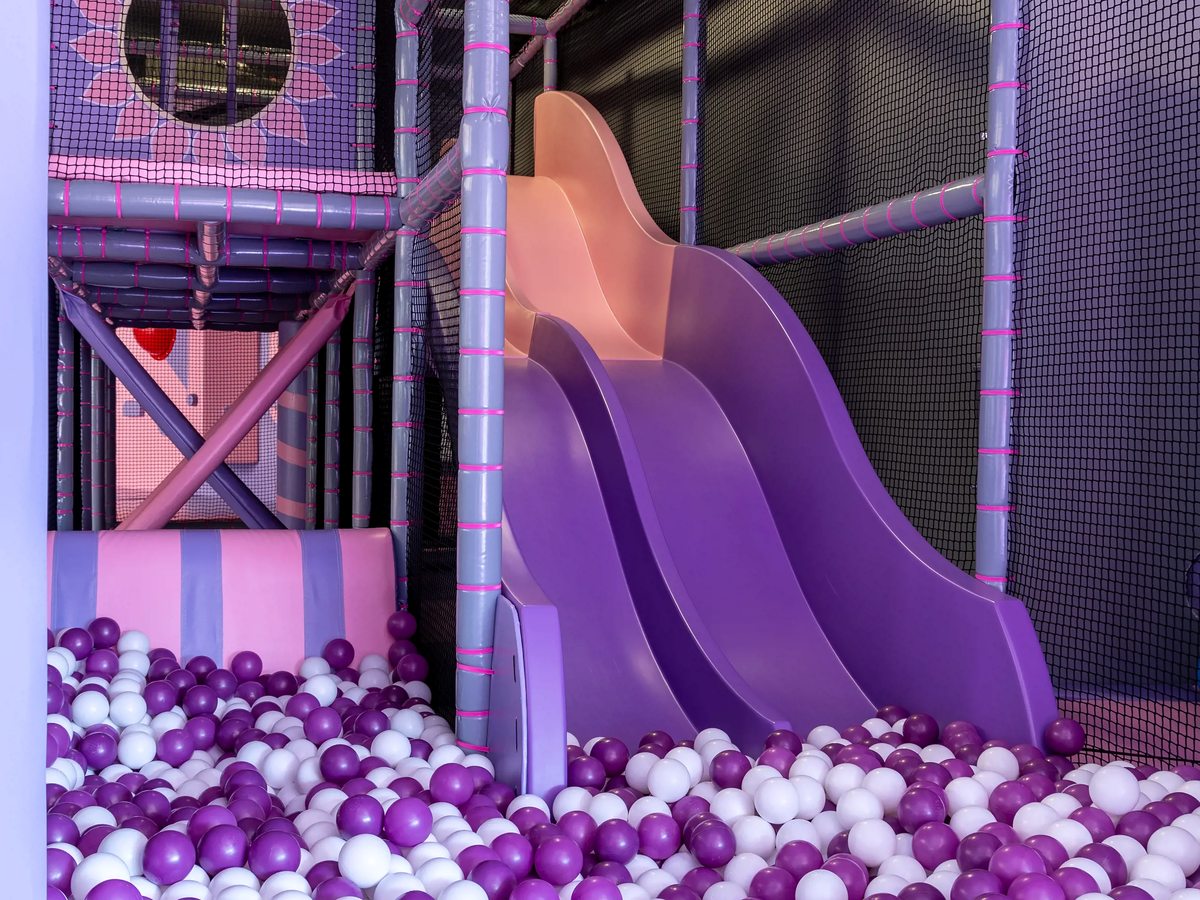 A purple twin slide running down into a ball pit of purple and white balls inside a netted soft-play frame.