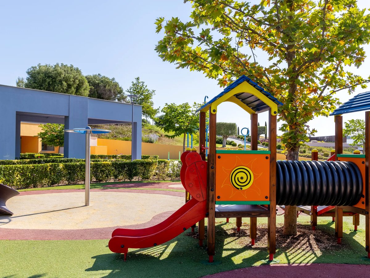 A wooden play frame with a red slide and a black crawl tunnel on soft coloured safety surfacing, shaded by a tree.